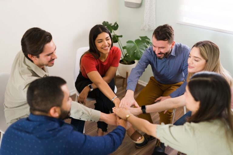A group of adults sitting in a circle during an ABA therapy support group meeting, smiling and joining hands in the center as a gesture of teamwork and encouragement.